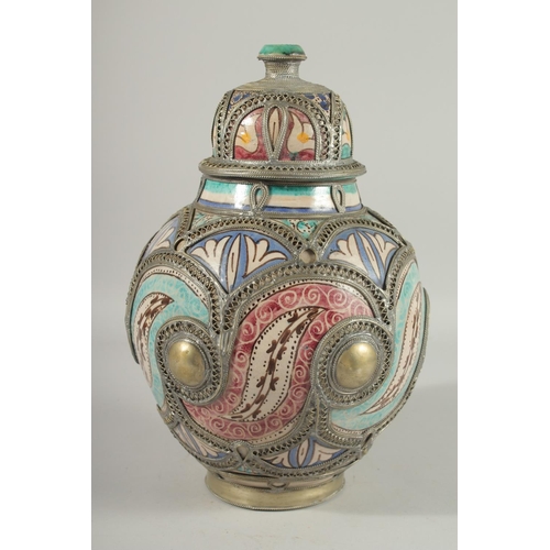 303 - A MOROCCAN METAL MOUNTED POTTERY LIDDED JAR, the jar painted with foliate motifs, 37cm high.