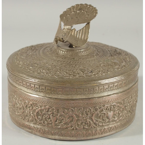 310 - A MALAYSIAN EMBOSSED SILVER CIRCULAR BOX, with figural peacock finial and foliate decoration, 18cm d... 