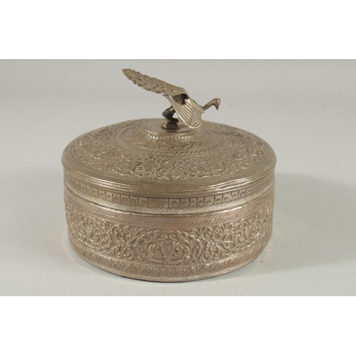 310 - A MALAYSIAN EMBOSSED SILVER CIRCULAR BOX, with figural peacock finial and foliate decoration, 18cm d... 