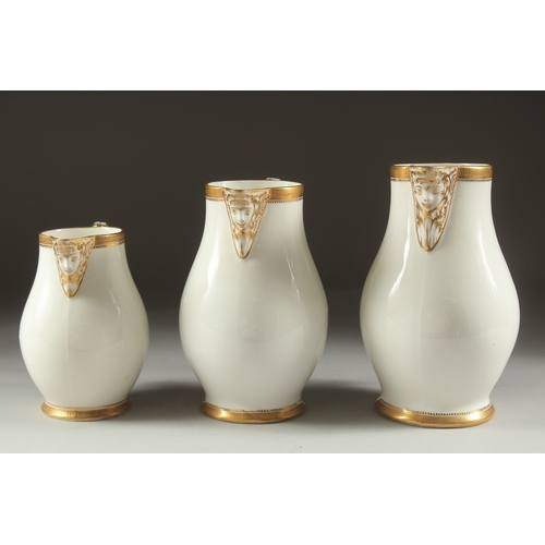 1074 - A GARNITURE OF THREE WHITE PORCELAIN GILDED JUGS with mask spouts.