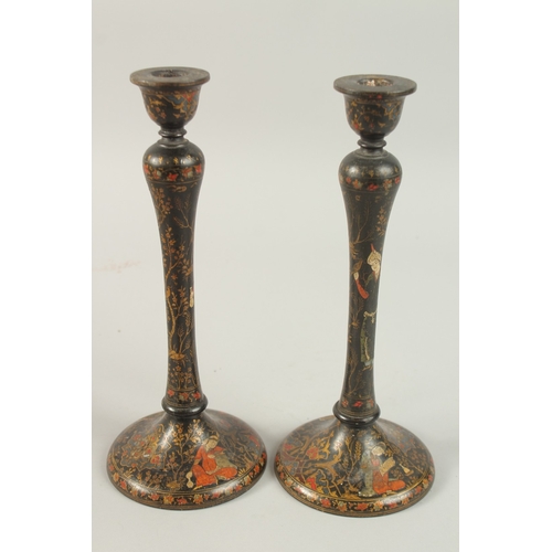 315 - A PAIR OF PERSIAN QAJAR LACQUERED WOOD CANDLESTICKS, decorated with figures and flora, 26.5cm high.