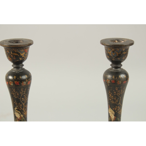 315 - A PAIR OF PERSIAN QAJAR LACQUERED WOOD CANDLESTICKS, decorated with figures and flora, 26.5cm high.