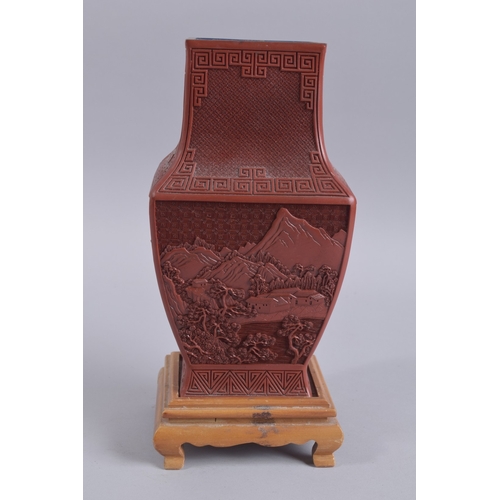 125 - A CHINESE RED CINNABAR LACQUER VASE ON WOODEN STAND, with landscape scenes and flora, 28cm high.