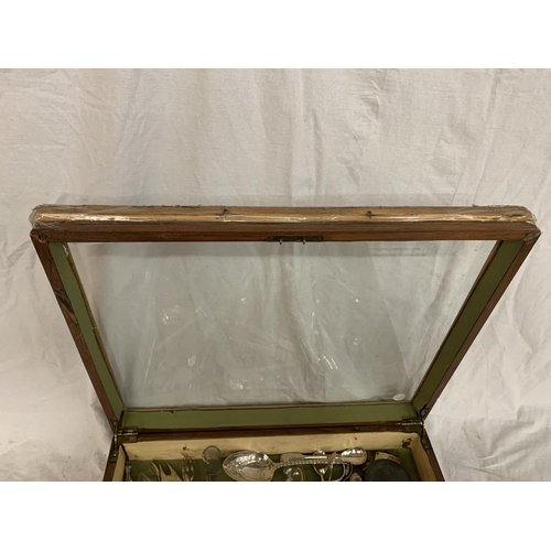 131 - A WOODEN FRAMED GLASS TOPPED JEWELLERY DISPLAY CABINET (A/F) WITH A LARGE QUANTITY OF EPNS