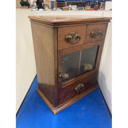 42 - A VINTAGE OAK SMOKERS CABINET WITH TWO SHORT UPPER DRAWERS, A GLAZED DOOR AND A LONG LOWER DRAWER AN... 