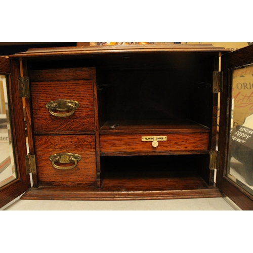 52 - AN OAK TWO GLASS DOOR SMOKERS CABINET WITH THREE INTERIOR DRAWERS ONE LABELED PLAYING CARDS COMPLETE... 