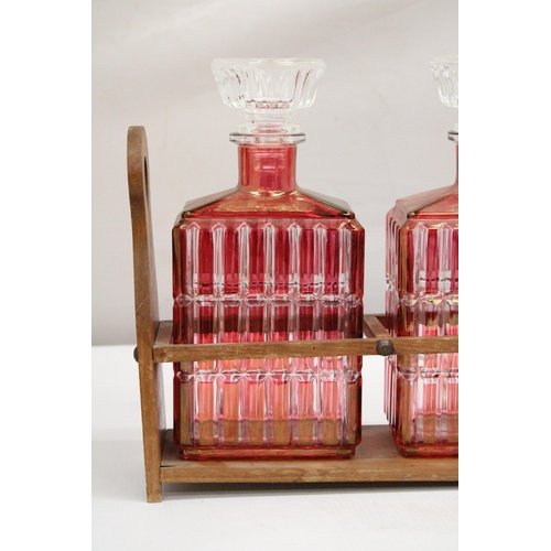 39 - A WOODEN HOLDER CONTAINING THREE AUSTRIAN STYLE 1930'S RUBY RED FLASHED ART DECO GLASS DECANTER LIQU... 