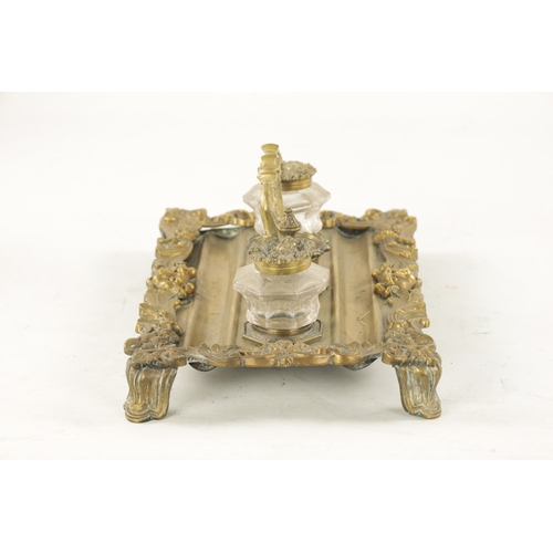 984 - A 19TH CENTURY BRASS ROCOCO INK STAND with glass ink bottles raised on a leaf cast and lions mask ba... 