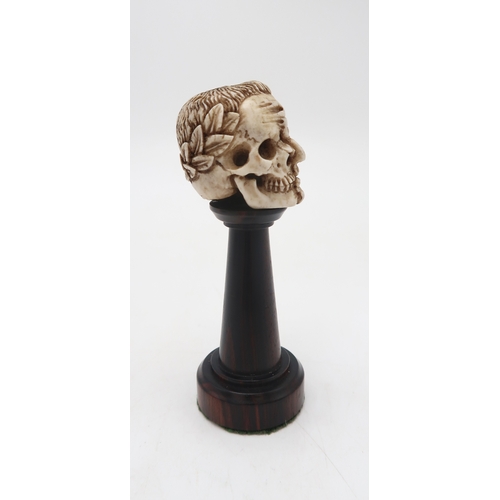 2254 - A CARVED BONE HALF SKULL/ HALF FACEwith a wreath of laurel to the back of the head, upon turned wood... 