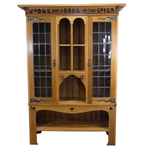 An Arts and Crafts oak motto Bookcase, in the manner of Liberty, having a flat solid oak overhanging cornice above the frieze which has an applied motto reading "Judge Not a Book by The Cover.", with twin rectangular Liberty style leaded glass doors and hand beaten strap work hinges which open to reveal two adjustable shelves, the base having an open central section with three pierced heart shaped motifs, typical of Liberty's furniture of this period, unmarked, W 122 cm x H 170 cm x D 43 cm. Provenance: property of a West Country Collector. After buying an Arts & Crafts house, the vendors researched and sought-out beautiful, often unique, Arts & Crafts pieces to furnish their home.