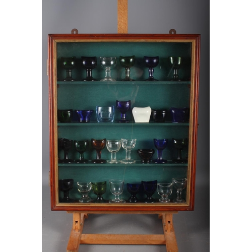 239 - A collection of twenty-nine 19th century and later glass and ceramic eye baths, in glass fronted dis... 