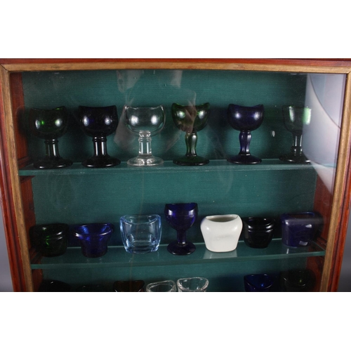 239 - A collection of twenty-nine 19th century and later glass and ceramic eye baths, in glass fronted dis... 