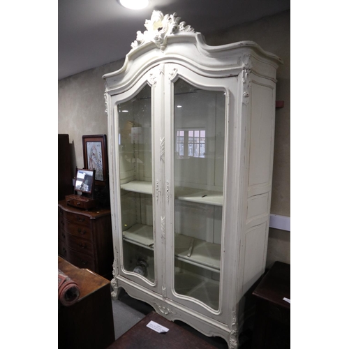 554 - A French carved and white painted armoire with scroll crest over two arch top glazed doors, on cabri... 