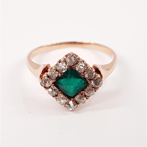 114 - AN EMERALD AND DIAMOND CLUSTER RING, with central emerald set at an angle, surrounded by a frame of ... 