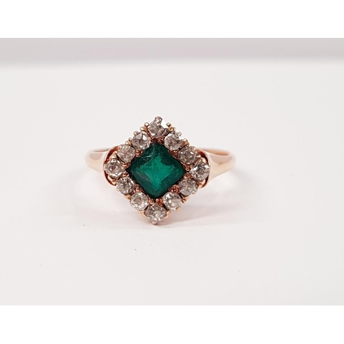 114 - AN EMERALD AND DIAMOND CLUSTER RING, with central emerald set at an angle, surrounded by a frame of ... 