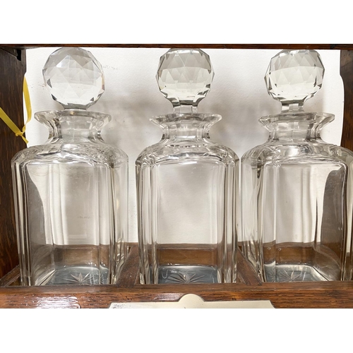 45 - AN ANTIQUE THREE BOTTLE TANTULUS, with three cut glass decanters/carafes, within oak frame with silv... 