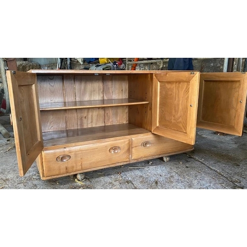 76 - AN ELM WOOD ERCOL THREE DOOR SIDEBOARD, with rectangular top above three doors opening to shelved in... 