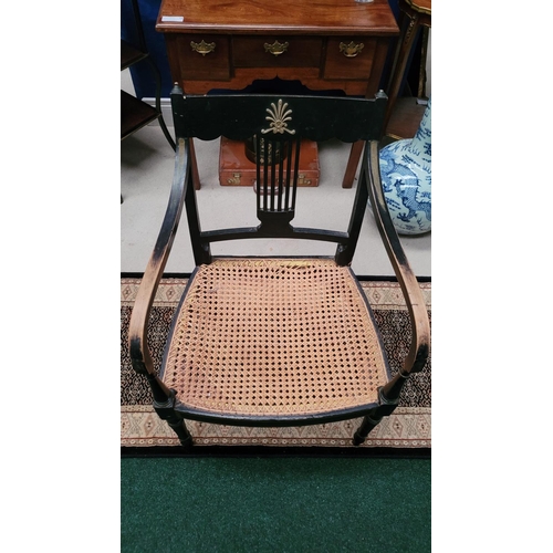 74 - A REGENCY EBONISED CANE-SEAT ARMCHAIR, with gilt mount detail to the curved crest rail and a lyre st... 