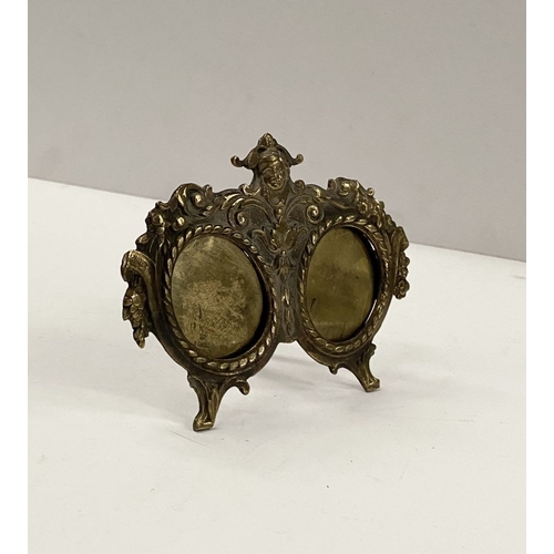 78 - A LATE 19TH CENTURY ORNATE BRASS DOUBLE PHOTO FRAME, the two oval framed with top border, figural mo... 