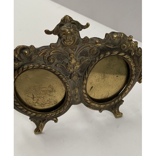 78 - A LATE 19TH CENTURY ORNATE BRASS DOUBLE PHOTO FRAME, the two oval framed with top border, figural mo... 