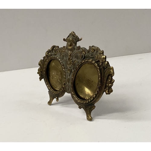 78 - A LATE 19TH CENTURY ORNATE BRASS DOUBLE PHOTO FRAME, the two oval framed with top border, figural mo... 