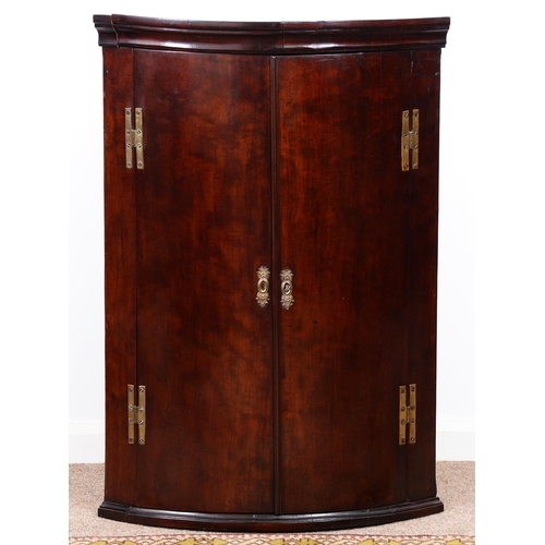 983 - A George III bow fronted mahogany hanging corner cupboard, fitted with shelves enclosed by figured d... 