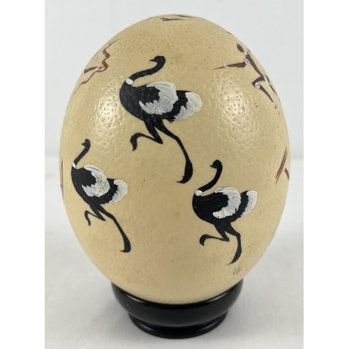 1262 - A hand painted ostrich egg decorated with an African hunting scene, on an ebony stand. Total height ... 