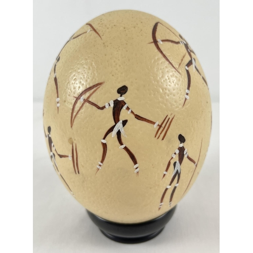 1262 - A hand painted ostrich egg decorated with an African hunting scene, on an ebony stand. Total height ... 