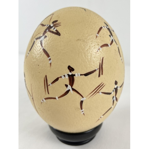 1262 - A hand painted ostrich egg decorated with an African hunting scene, on an ebony stand. Total height ... 