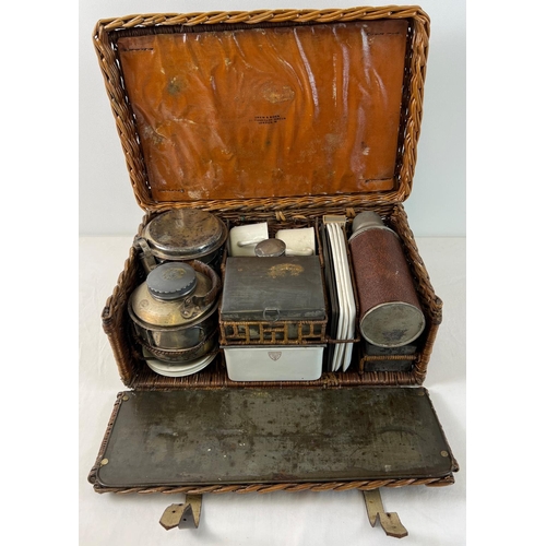 An early 20th century Drew & Sons 'En Route' picnic hamper for 4, circa 1910. With leather straps and handle. Named to basket interior lid and most of the pieces. Pull down front tray with metal lined work station. Hamper includes: Pot stand with decorative pierced panel; burner with adjustable wick; fuel canister, saucepan & lid with fold out handles; kettle with screw lid & spout and wicker handle; 4 ceramic saucers; 4 teacups (only 1 original); rectangular steel canister with ceramic liner; 3 ceramic picnic trays (1 missing); thermos flask, glass bottle with screw lid and 2 small steel lidded canisters.