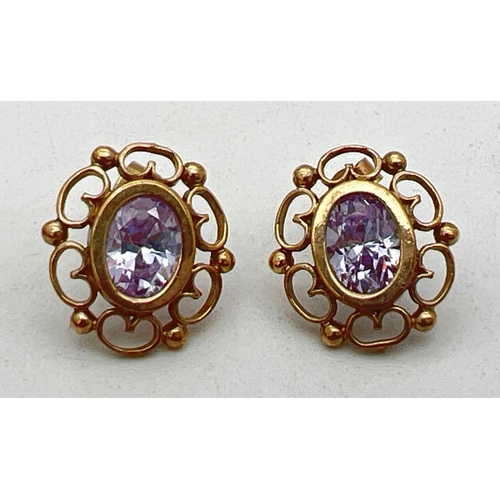 243 - A pair of yellow metal stud style earrings with scroll design mount set with an oval cut pale lilac ... 