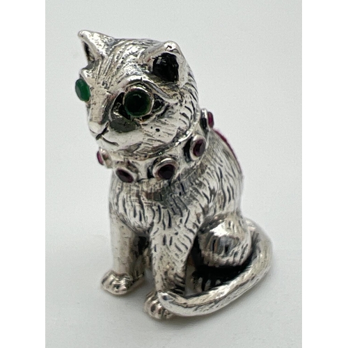 1123 - A small silver pin cushion modelled as a cat with green stone set eyes and red stone set collar. Sta... 