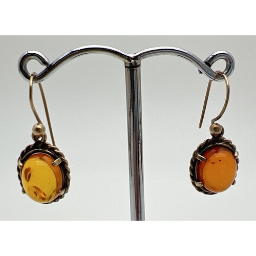 1119 - A pair of yellow metal amber set drop style earrings. Oval cut amber cabochon in a rope detail mount... 