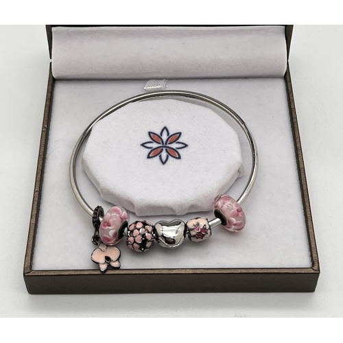 1063 - A boxed costume jewellery bangle with 5 charms by Khloelle Charms. 3 charms with pale pink enamel de... 