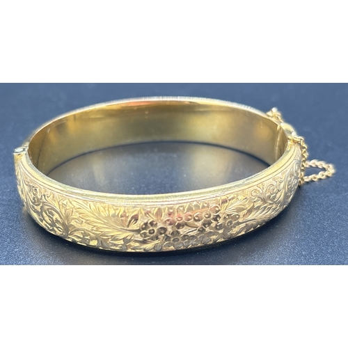 2 - A 9ct gold hinged bangle with half floral engraved decoration. Push release clasp with safety chain.... 