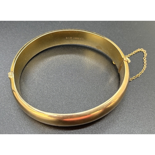 2 - A 9ct gold hinged bangle with half floral engraved decoration. Push release clasp with safety chain.... 