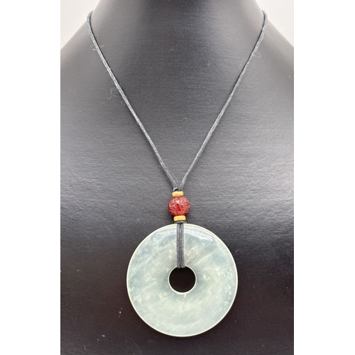 1081 - A circular jade pendant necklace with carved carnelian bead on an adjustable black leather thong. Pe... 