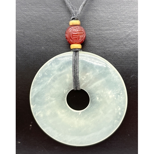 1081 - A circular jade pendant necklace with carved carnelian bead on an adjustable black leather thong. Pe... 