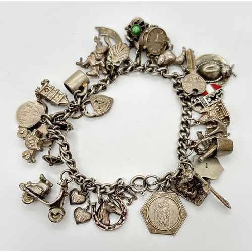 A vintage white metal charm bracelet with 28 silver and white metal charms, spring ring clasp & safety chain. Charms include clock, cowboy hat, enameled shields, scooter, throne, cutlass, bucket and a flying carpet. Total weight approx. 64.9g.