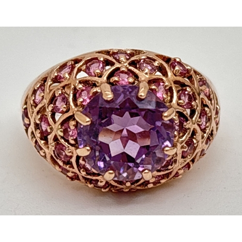 A 9ct gold, amethyst and pink tourmaline ring in a dome style with pierced design. A central raised claw set round amethyst, surrounded by 30 round cut pink tourmaline stones set in a lattice design to both shoulders and sides of mount. Fully hallmarked inside band. Ring size P. Total weight approx. 5.7g.
