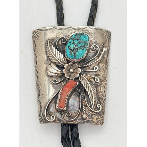A vintage sterling silver Navajo Native American bolo tie by Sheryl Martinez. Tie clasp in a floral design set with a piece of turquoise and coral, with hammered decoration to edges. 19" black leather plaited cord and sterling silver aglets. Markers initials and Sterling stamped to back of clasp. Approx. 5 x 4.5cm.
