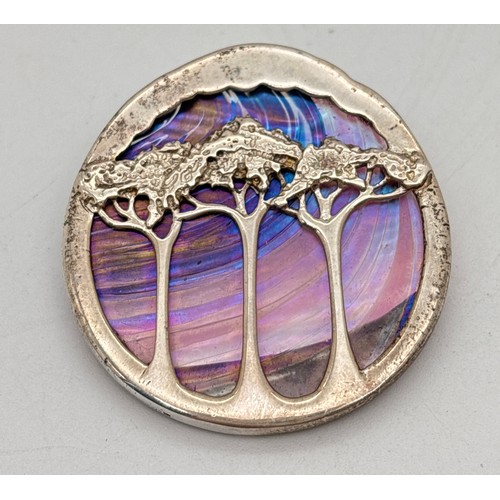 A vintage Pat Cheney/John Ditchfield silver and iridescent glass pendant brooch. Depicting a night sky behind Art Nouveau stylised trees. Glass panel in purple and blue tones. Pat Cheney and Scottish silver marks to side of mount. Fixed bale and brooch pin to back. Approx. 4cm diameter.