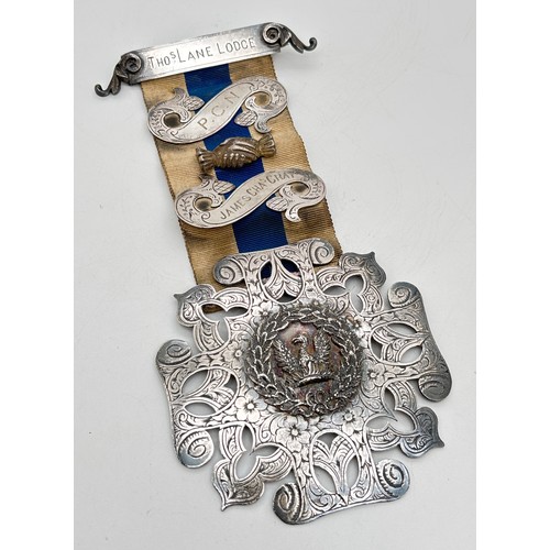 A large Victorian silver Masonic jewel awarded for services, on a blue and white ribbon. A top bar with foliate decoration engraved "THos Lane Lodge", with 2 scroll sections featuring acorn decoration to centre of the ribbon engraved "P.C.N. and James Chars Gray", a handshake applique (Not silver) between each scroll. A large square pierced work medallion to the bottom of the ribbon with Phoenix and laurel wreath detail to centre and foliate engraving throughout.  Back of medallion inscribed "Presented by the Officers & Bros of the Thos Lane Lodge to PCN Bro James Chas Gray as a mark of respect for past services Dec.20.1882". Silver marks to top bar and scrolls, full hallmarks to medallion for Richard Britton London 1882. Total length approx. 16.5cm. Total weight approx. 85.4g.