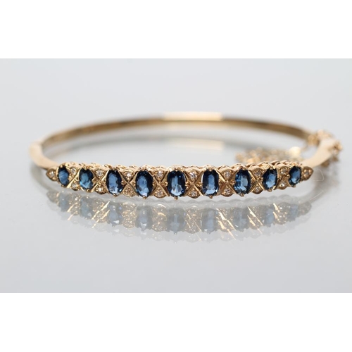165 - A SAPPHIRE AND DIAMOND STIFF HINGED BANGLE, the upper section grain set with nine oval facet cut sap... 