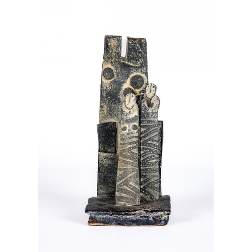 81 - JOHN MALTBY (1936-2000) - a matt grey stoneware sculpture of two figures standing before a cut slab ... 