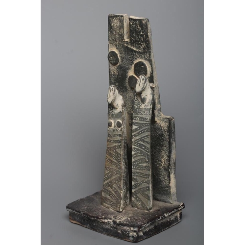 81 - JOHN MALTBY (1936-2000) - a matt grey stoneware sculpture of two figures standing before a cut slab ... 