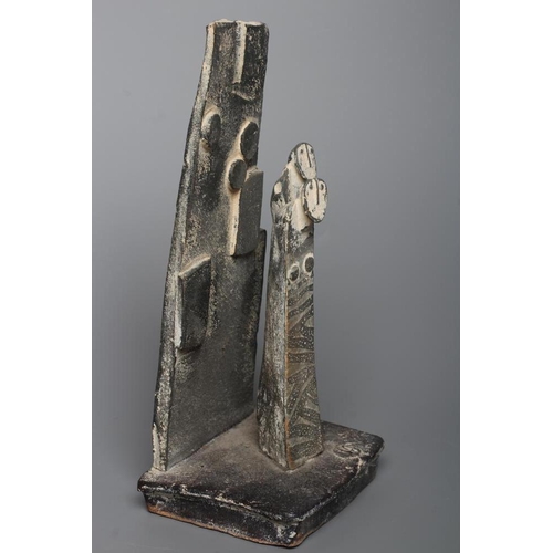 81 - JOHN MALTBY (1936-2000) - a matt grey stoneware sculpture of two figures standing before a cut slab ... 