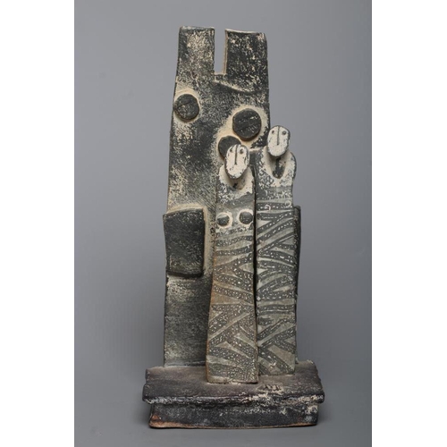 81 - JOHN MALTBY (1936-2000) - a matt grey stoneware sculpture of two figures standing before a cut slab ... 