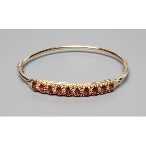 305 - A STIFF HINGED BANGLE, the upper section claw set with ten oval facet cut garnets, indistinctly stam... 