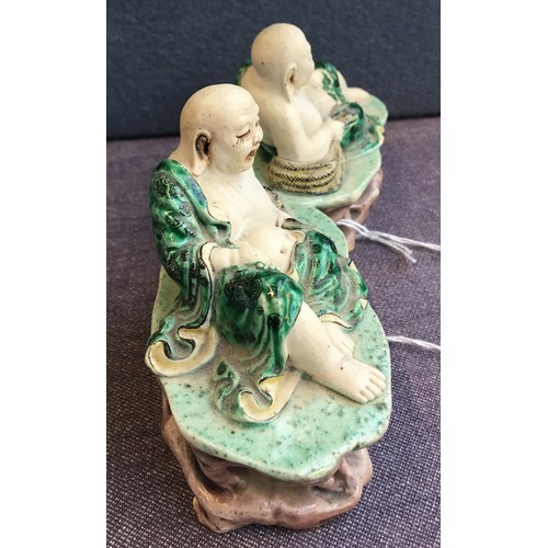 169 - A pair of famille verte biscuit porcelain Budai figures, with green underglaze robes, reclined upon ... 